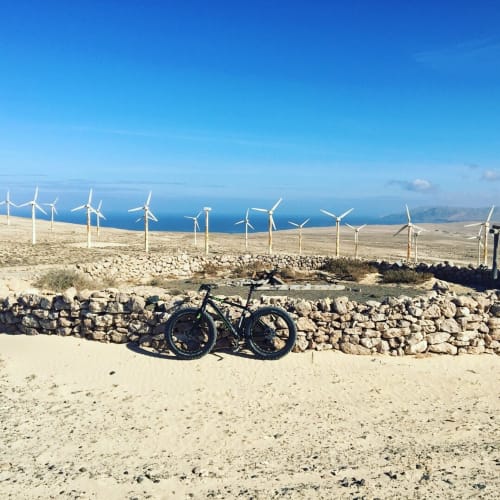 Bicicleta apoyada en muro de piedra con aerogeneradores y mar azul en Smart Rent Fuerteventura