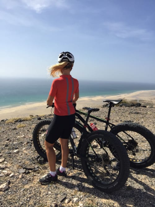 Ciclista con jersey rojo y casco contemplando la playa de Fuerteventura y el océano desde un acantilado costero