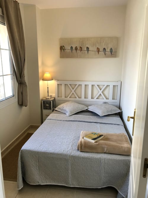 Luminosa habitación del apartamento Smart Rent Fuerteventura con ropa de cama azul a rayas, cabecero blanco, lámpara de noche y pájaro