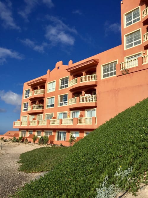 Edificio de apartamentos en la playa de color coral en Fuerteventura con barandillas blancas, múltiples balcones y vegetación desértica
