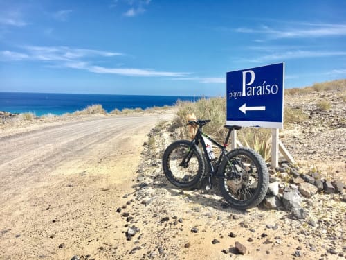 Bicicleta de montaña apoyada en señal de Playa Paraíso en Fuerteventura con océano turquesa y costa arenosa visible.