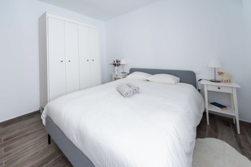 Dormitorio moderno con cama gris, ropa de cama blanca, armario blanco y mesita de noche en apartamento Smart Rent Fuerteventura