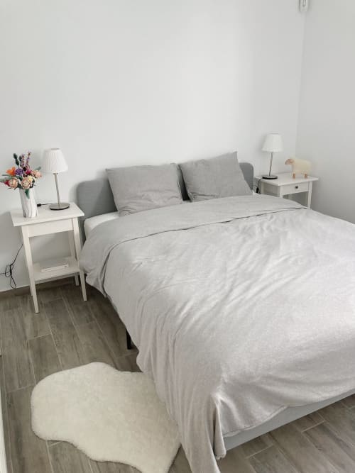 Dormitorio moderno en Smart Rent Fuerteventura con cama tapizada gris, mesitas blancas, lámpara de mesa, flores frescas y