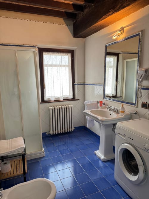 Spacious bathroom with blue tile floor, white pedestal sink, shower, washing machine, and exposed wooden beams at Agriturismo