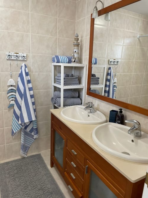 Baño náutico con doble lavabo, espejo, tocador de madera, estantería blanca con toallas rayadas y paredes de azulejos beige en Smart