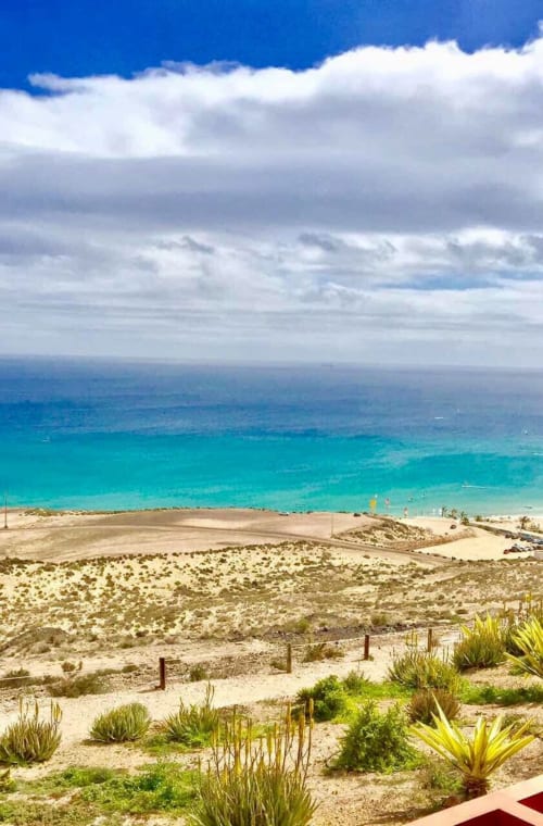 Vista de océano turquesa y playa de arena desde apartamento en Fuerteventura con paisaje desértico y palmeras