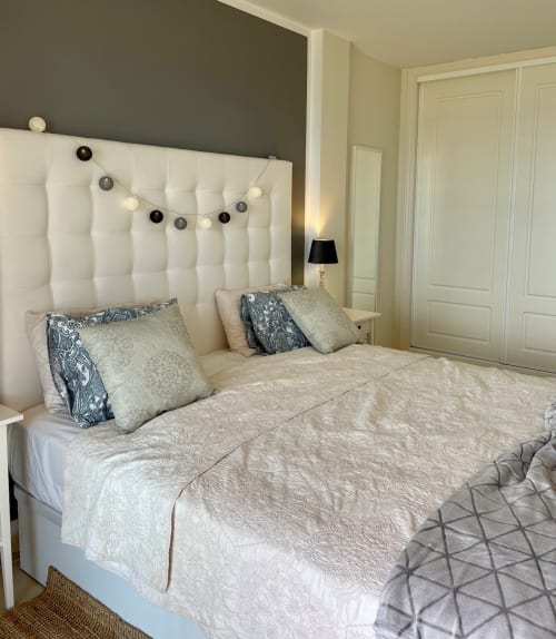 Dormitorio moderno con cabecera blanca capitonada, ropa de cama beige, cojines decorativos y luces LED en Lituanica PlayaParaiso