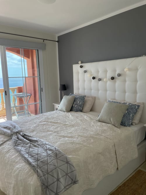 Dormitorio moderno en Smart Rent Fuerteventura con cabecero tapizado blanco, balcón con vista al océano y pared acentuada gris