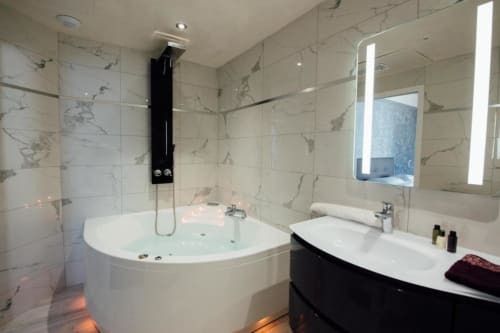 Salle de bains moderne avec carrelage en marbre, baignoire d'angle spa, double vasque et miroirs éclairés à l'Hôtel Amirauté La Baule