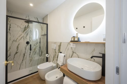 Salle de bains moderne du Palazzo Garibaldi Parma avec carrelage marbre, miroir rond éclairant, lavabo blanc, toilettes et douche à l'italienne