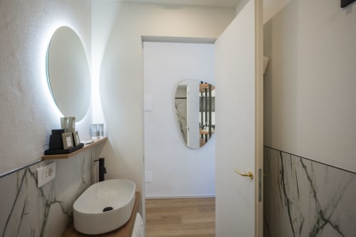 Salle de bains moderne du Palazzo Garibaldi Parma avec miroir ovale illuminé, lavabo blanc et revêtement mural effet marbre.