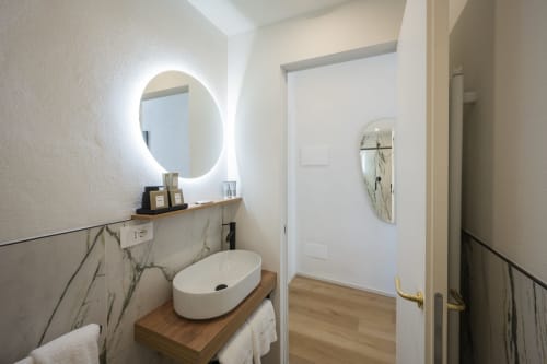Salle de bain moderne au Palazzo Garibaldi Parma avec miroir rond illuminé, lavabo blanc et accents muraux effet marbre