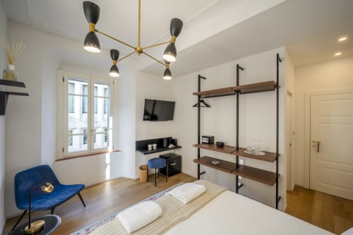 Chambre moderne au Palazzo Garibaldi Parma avec lit, fauteuil bleu, étagères en bois, suspensions et TV.