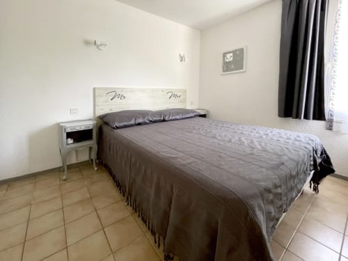 Chambre lumineuse de l'appartement LOCADIGNE avec lit queen-size, tête de lit blanchie, table de nuit, carrelage et fenêtre