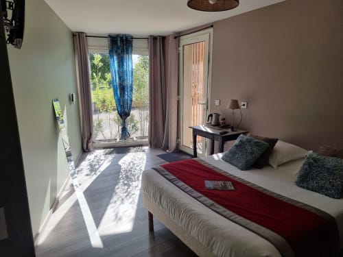 Chambre lumineuse de l'Hôtel Amadour à Rocamadour avec lit queen, accès à la terrasse, bureau et vue sur le jardin par les portes vitrées.