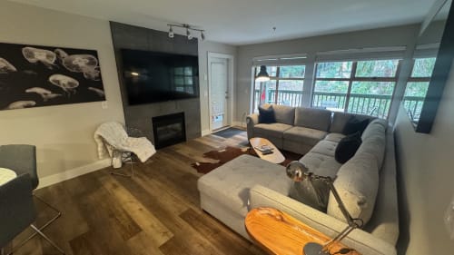 Modern Whistler apartment living room with L-shaped gray sofa, fireplace, TV, hardwood floors, and large windows overlooking