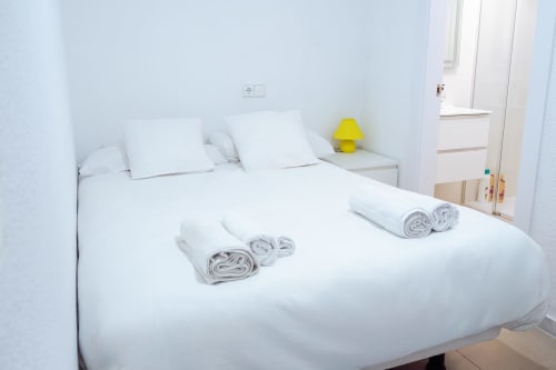 Habitación blanca y limpia en Guest Homes Málaga con toallas enrolladas en la cama y lámpara amarilla en la mesita de noche