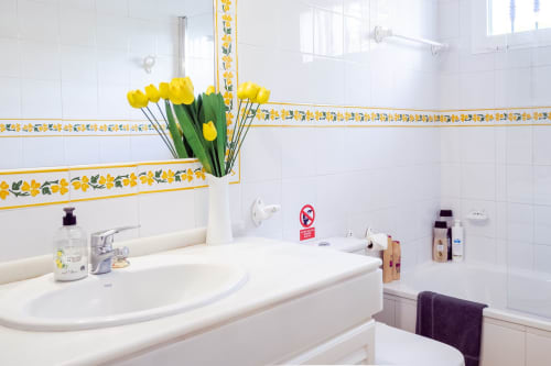Baño luminoso en Guest Homes Málaga con azulejos blancos, cenefa floral amarilla y tulipanes amarillos en florero junto al lavabo