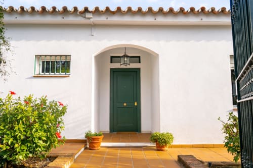 Entrada con puerta arqueada verde en Guest Homes Málaga, muros blancos, azulejos de terracota y plantas en macetas