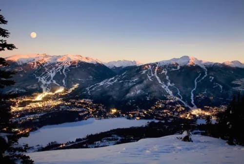 Whistler mountain resort at twilight with snow-covered peaks, illuminated valleys, lake, and full moon overhead