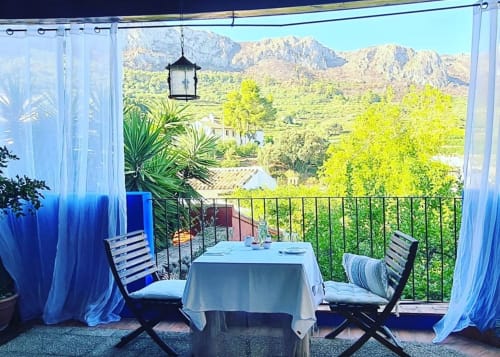 Shaded terrace at Casa Gallinera vacation home in Benissivà with mountain views, dining table, and blue curtains