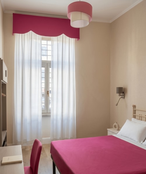 Camera Matrimoniale Economy dell'Hotel Principe Amedeo con letto rosa, accenti bordeaux, tende bianche e vista sulla città