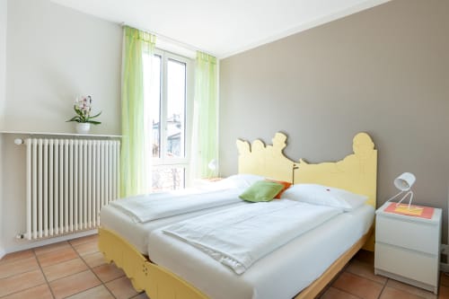 Camera luminosa presso la Residenza Motta Locarno con letto a struttura gialla, biancheria bianca, tende verdi e vista sul giardino dal balcone