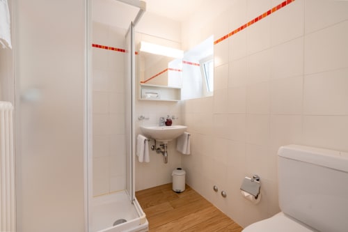 Salle de bain moderne de l'appartement Residenza Motta à Locarno, avec douche, lavabo, toilettes, sol en bois et accents en carrelage rouge