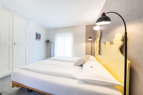 Camera da letto luminosa presso Residenza Motta a Locarno con testiera gialla, biancheria bianca, lampada ad arco e vista sul giardino