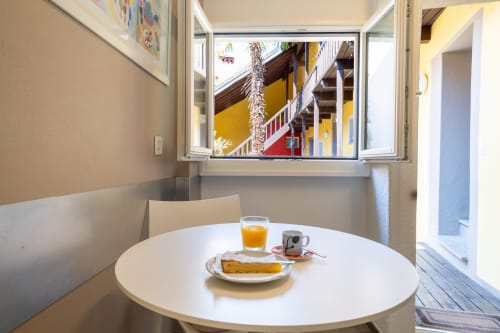 Coin petit-déjeuner à l'appartement Residenza Motta Locarno avec table ronde, jus de fruit, viennoiserie et vue sur le jardin coloré par la fenêtre