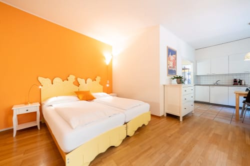 Luminoso monolocale alla Residenza Motta a Locarno con parete arancione, letto giallo, cucina bianca e pavimenti in legno.