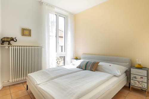 Camera luminosa presso la Residenza Motta a Locarno con pareti gialle, letto bianco, radiatore e porte-finestre con vista sulla città