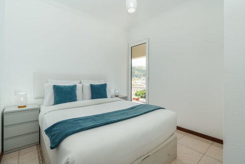 Bright modern bedroom with white walls, teal accents, and sea view balcony at Azul Infinito Casa, Arco da Calheta