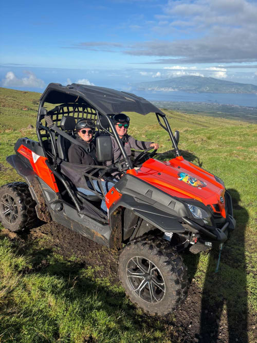 Two guests in red UTV on green hillside with coastal views near Adega da Quinta, Madalena