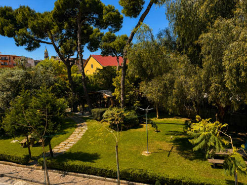 Rigoglioso giardino di Palazzo Sebeto a Napoli con alberi adulti, prato curato e cappella gialla con tetto rosso sullo sfondo