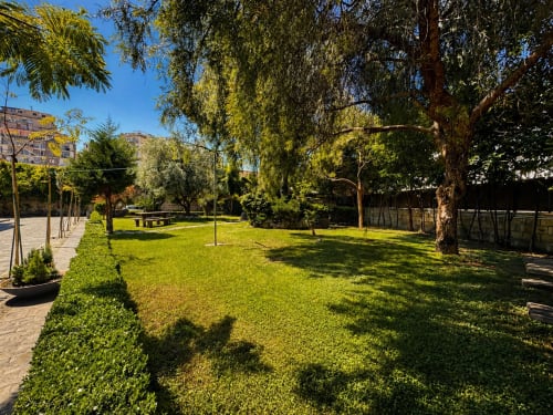 Palazzo Sebeto Napoli: giardino lussureggiante con alberi maturi, prato curato e vialetti pavimentati sotto cielo azzurro