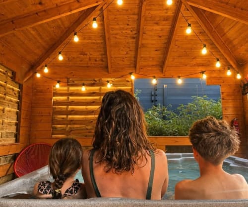 Trois hôtes profitent d'un bain à remous dans un lodge en bois aux Gites D'armor, Treguidel, avec guirlandes lumineuses et vue montagne en soirée