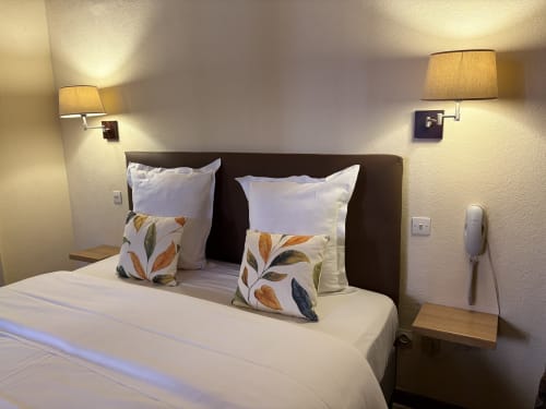 Chambre d'hôtel moderne à l'Hôtel de l'Ange avec draps blancs, coussins décorés de feuilles et lampes de lecture murales