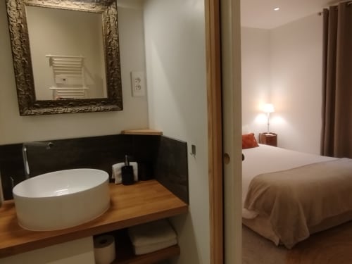 Salle de bain attenante moderne avec vasque blanche et meuble en bois, ouvrant sur la chambre de l'appartement Villa Voltaire