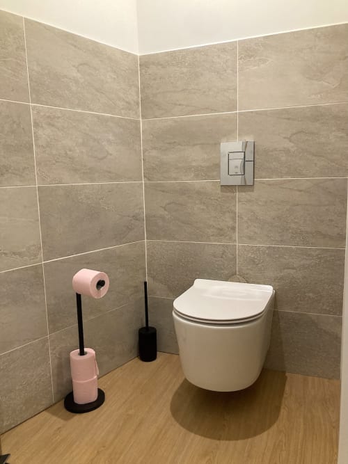 Baño moderno con inodoro blanco montado en pared, azulejos beige, piso de madera y dispensador de papel rosa en Au Château De