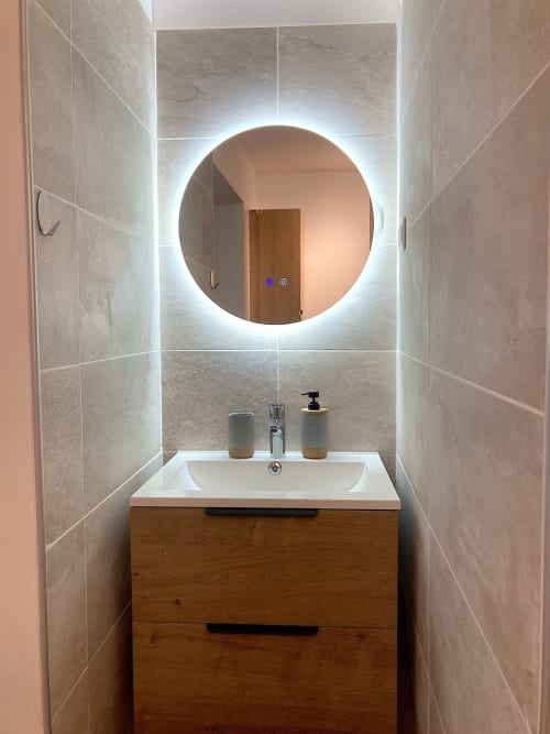 Baño moderno con espejo redondo iluminado, lavamanos blanco, mueble de madera y paredes de mármol en Au Château De Villelongue