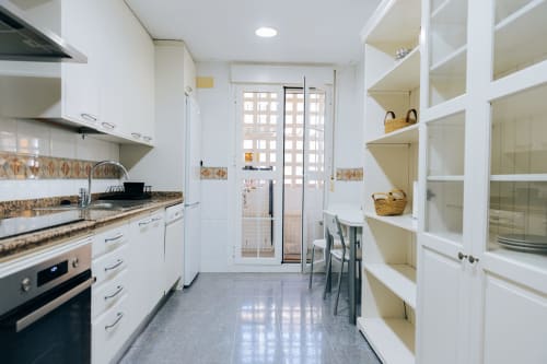 Modern white kitchen in Itaca by BH Marbella with granite counters, stainless appliances, open shelving, and balcony doors