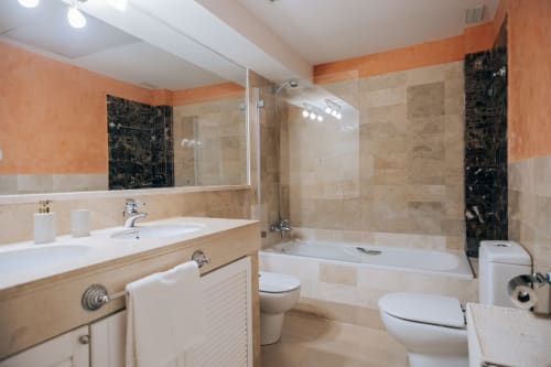 Modern bathroom at Rodas by BH Marbella with dual vanity, glass shower enclosure, and elegant travertine tiling