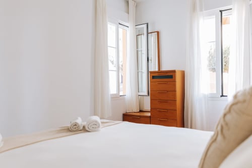 Bright bedroom at Rodas by BH Marbella with white linens, wooden dresser, mirrors, and garden view through French doors