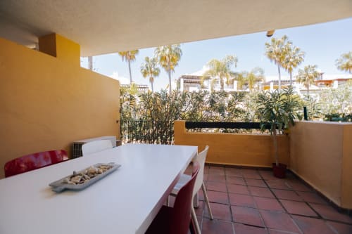 Marbella apartment terrace with white dining table, red chairs, and palm tree garden views