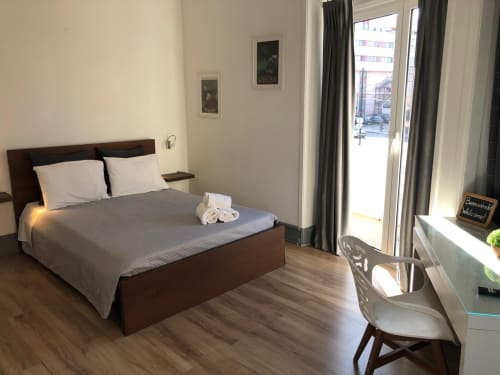 Quarto moderno com cama de madeira escura, almofadas brancas, roupa de cama cinzenta, secretária com cadeira branca e terraço com vista para a rua de Coimbra