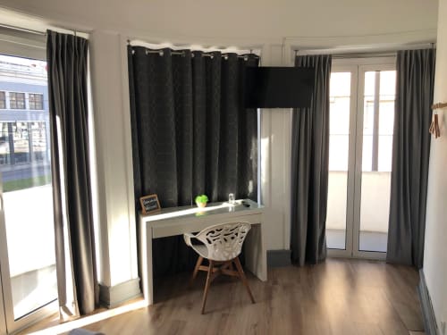 Quarto de hotel moderno com secretária, cadeira, televisão e janelas do chão ao teto com cortinas cinzentas com vista para Coimbra