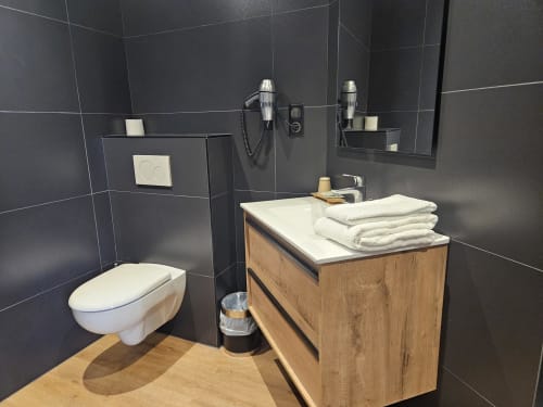 Salle de bains moderne avec carrelage noir, toilettes blanches, meuble de salle de bains en bois avec lavabo et serviettes blanches à l'Hôtel La Route d'Argent
