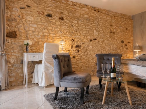 Chambre élégante à La Biche Au Bois avec murs en pierre apparente, fauteuil gris capitonné et plateau de champagne à Moncléra