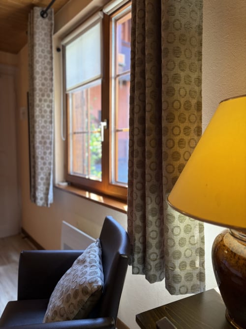 Chambre confortable à l'Hôtel de l'Ange à Niedermorschwihr avec fauteuil, rideaux à motifs et vue sur la fenêtre